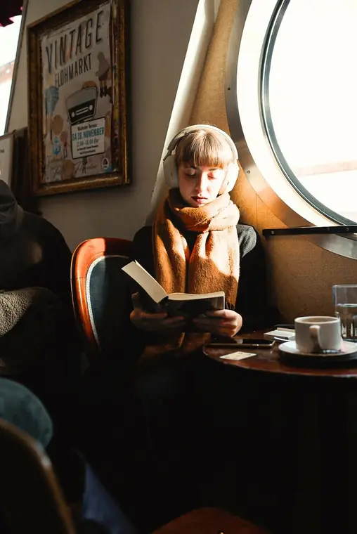 Frau mit Kopfhörern liest ein Buch am runden Fenster eines Cafés