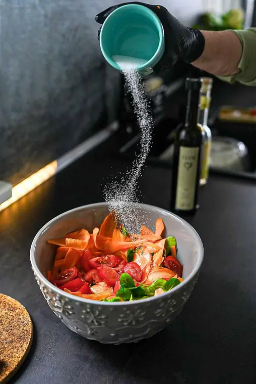 Salz rieselt aus einer Schale auf einen Salat aus Tomaten und Karotten